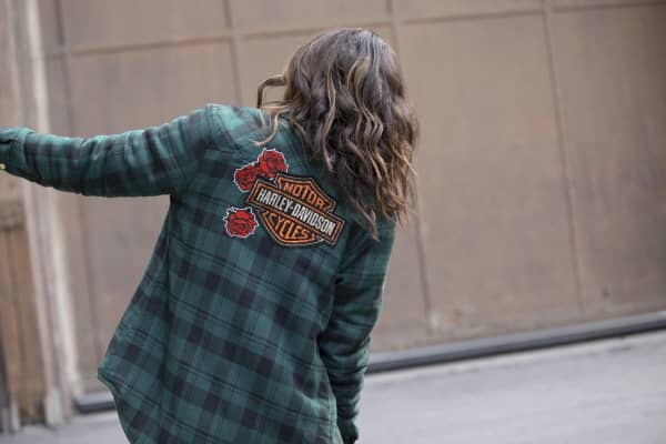 A woman facing away with long, brown, wavy hair leans to the right wearing a green plaid Harley-Davidson long-sleeve flannel shirt from the Harley-Davidson Garage collection.
