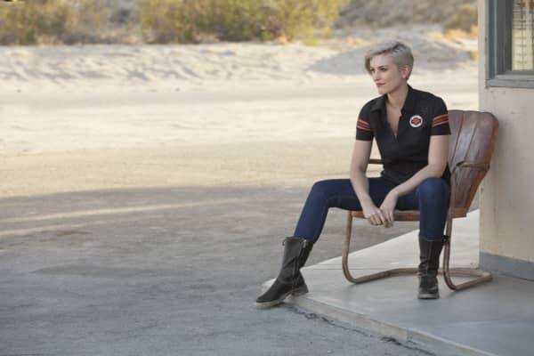 A woman with short blond hair sitting in a chair wearing a black V-neck polo from the Harley-Davidson Genuine MotorClothes collection.
